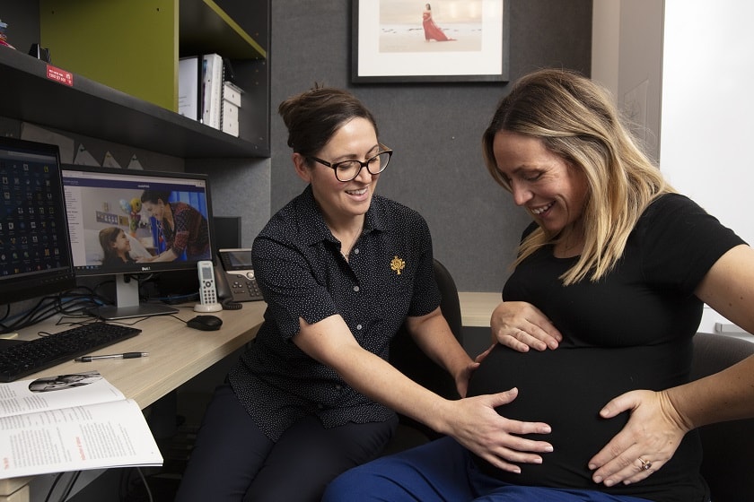 Caregiver putting hands on a pregnant and smiling patient's abdomen in a consulting room.
