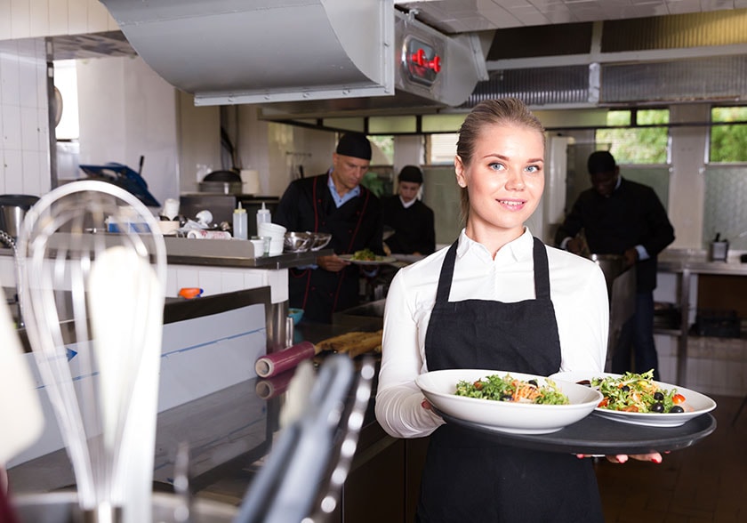 Person in a commercial kitchen holding a serving tray with salads, other colleagues in the background preparing food