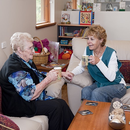Volunteer wearing uniformed vest sits with senior woman in lounge area drinking tea together