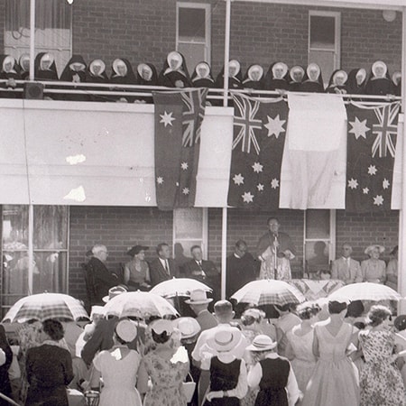 Black and white image from the opening of St Joseph’s Private Hospital Bicton on 11 November 1956 where a crowd watches nuns on a balcony with Australian flags draped over the balustrade