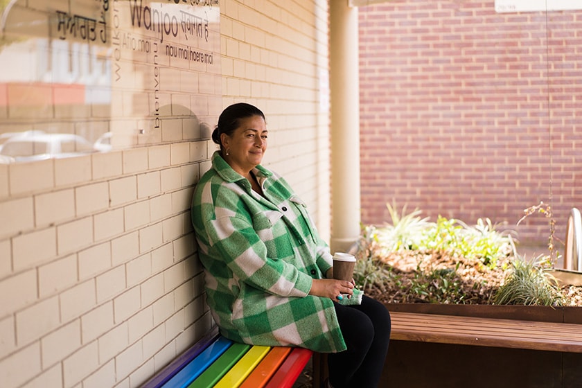 Person sitting in a commercial verandah setting on a wooden bench chair painted in rainbow stripes, holding a takeaway cup and looking out pondering