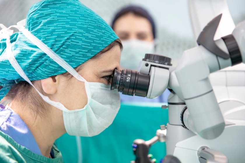 A surgeon in scrubs, mask, and cap closely examines a surgical microscope during a procedure, with another masked medical professional blurred in the background.