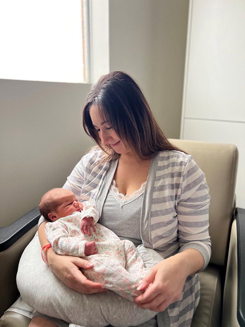 Amanda Mercer (Mother) sits in a chair cradling her swaddled baby.
