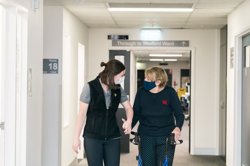 Caregiver holding hands with a client who is walking down the ward corridor using a walking frame and looking at one another