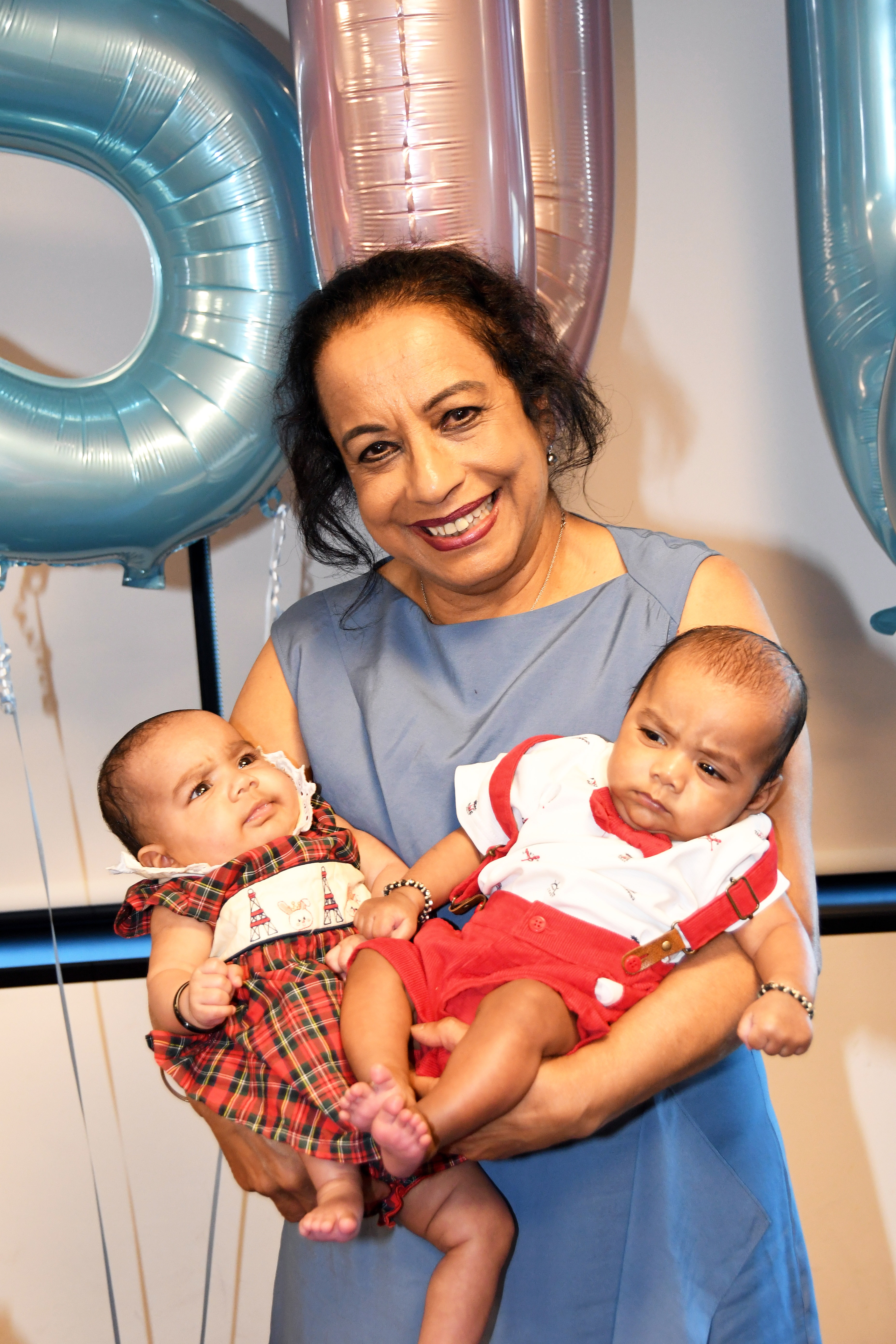 A woman in a blue dress holds 2 baby twins in front of pink and blue balloons