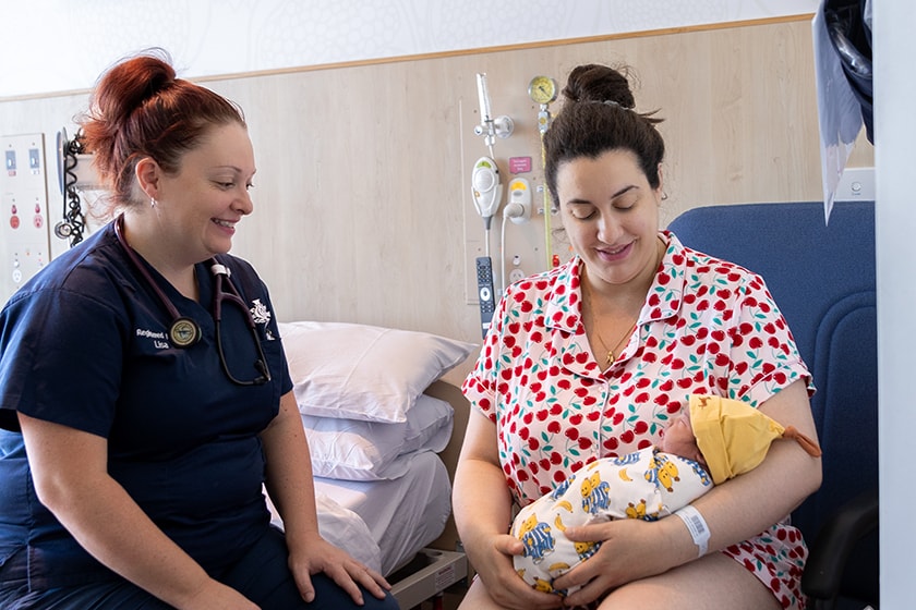 A person sits in a chair holding a newborn baby whilst a caregiver sits on the bed beside them looking at the baby.