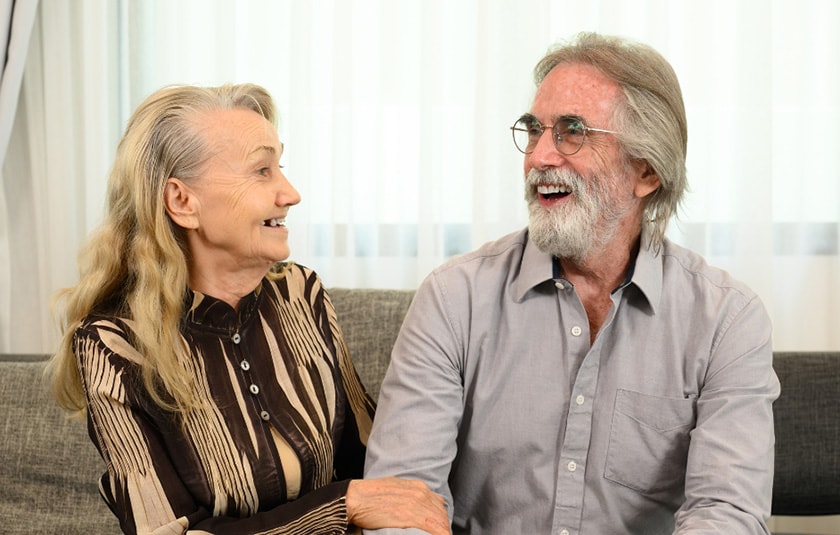 Two elderly people sit on a couch together laughing.