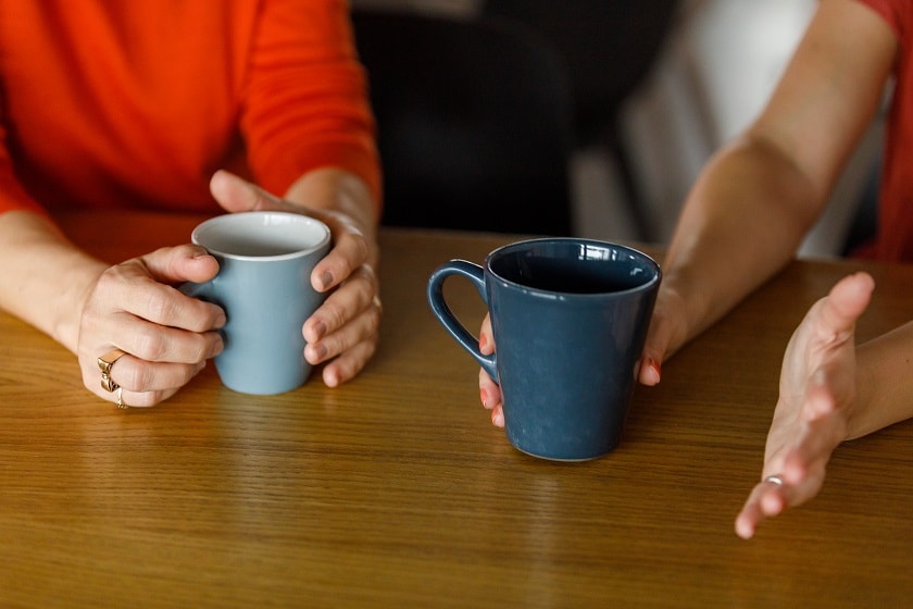 Two people holdings coffee cups sitting at a table.