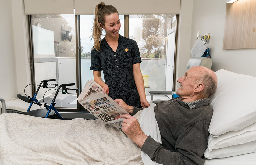 Caregiver at bedside smiling with a patient who is reading a newspaper