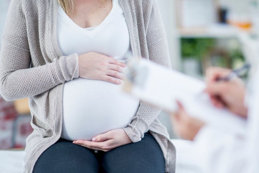 A person holding their pregnant belly while a person stands in the foreground writing on a clipboard.