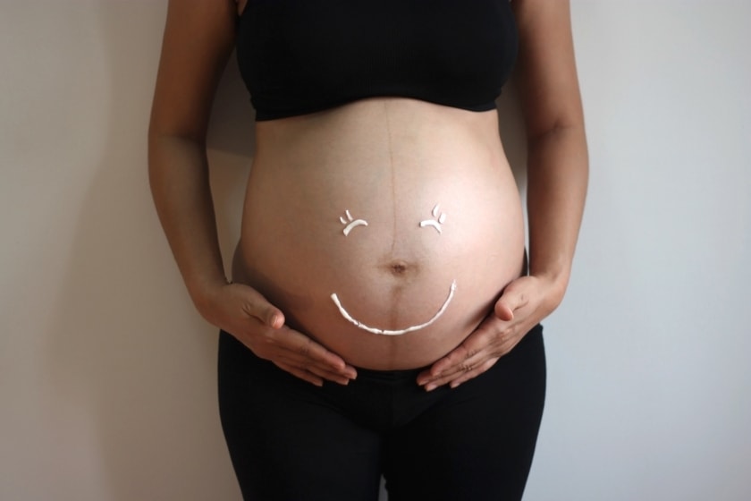 A women shows her pregnant belly with her hands folded underneath her belly, a smiley has been drawn on her belly with the bellybutton as the nose.