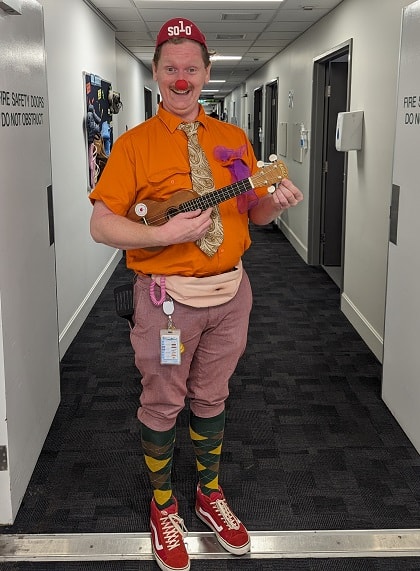 A person dressed in a bright orange shirt wearing a red nose, high socks and holding a ukulele.