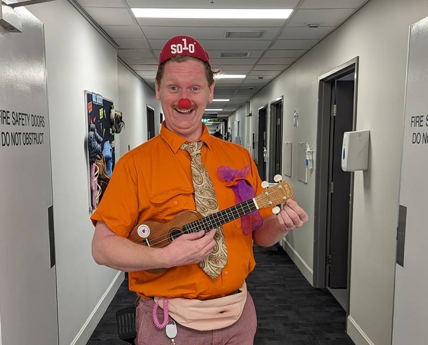 A person dressed in a bright orange shirt wearing a red nose and holding a ukulele.