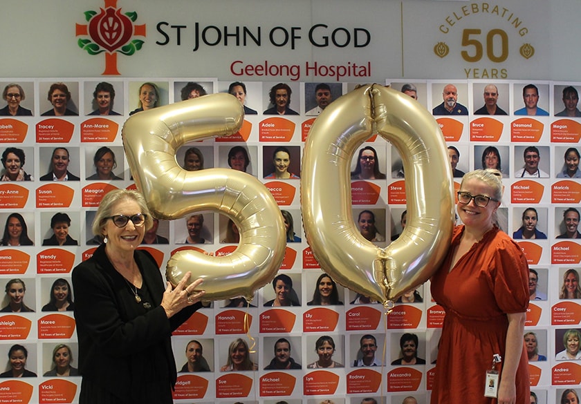 Two caregivers holding balloons shaped as the numbers five and zero stand in front of a mural of caregivers head shot photos.