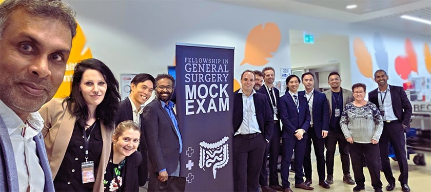 A line of people standing around a sign that says Fellowship in General Surgery Mock Exam
