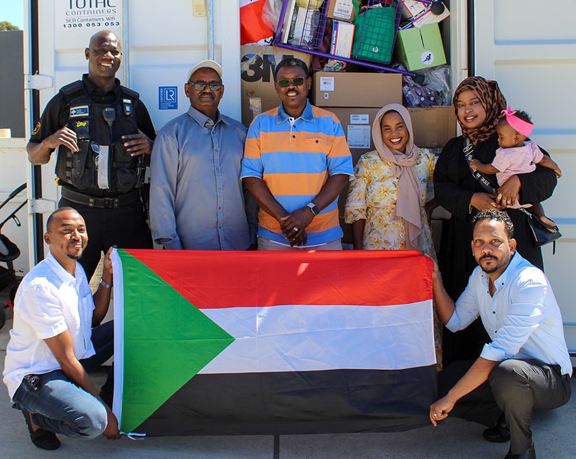 Hospital Security Team Leader, Saeed Saeed and members of the Labayka Ya Watani humanitarian group holding a Sudanese flag standing in front of a packed shipping container.