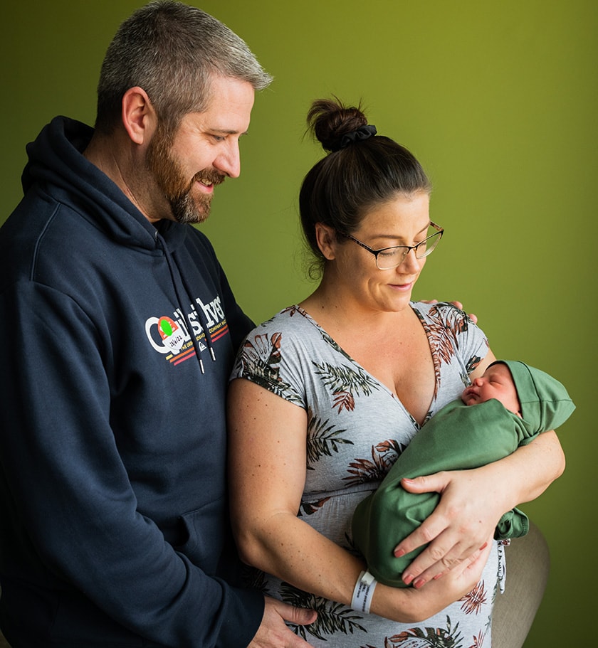 Maternity patient and partner looking at newborn baby being crading in mother's arms swadled in a green wrap and wearing a beanie