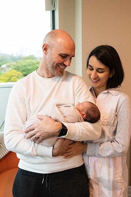 Mother and father standing in patient room with the father cradling a newborn baby in his arms whilst they both look down at the baby, grinning.