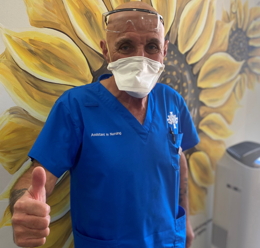 St John of God Mt Lawley Hospital Assistants in Nursing Lee wearing scrubs and a face mask giving a big thumbs up in front of a wall with a large sunflower print