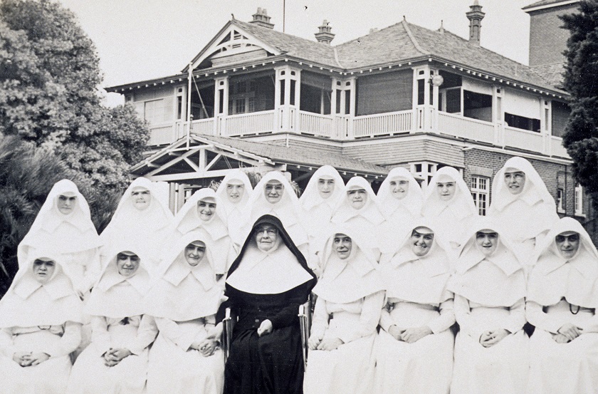17 Mercy Sisters in white habits seated in two rows with Mother Superior in a black habit. Behind the women is a two story heritage listed house.  