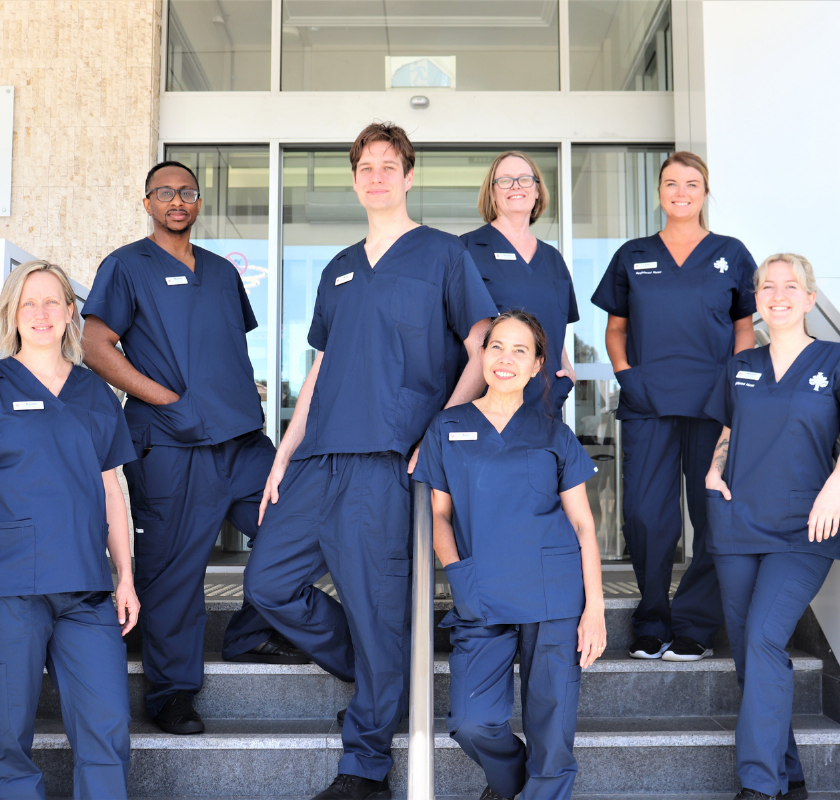Seven nursing caregivers standing at the front entrance stairs of St John of God Mt Lawley Hospital wearing new navy blue scrub uniforms. 