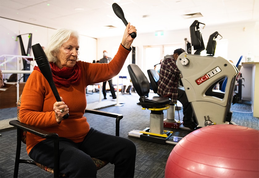 Person sitting on a chair in a rehabilitation gymnasium undergoing an exercise holding a baton shaped instrument in each hand