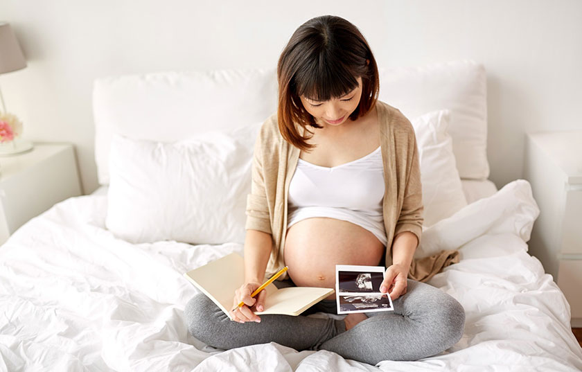 Heavily pregnant person sitting cross legged on a bed covered in white linens looking down at images from an ultrasound and writing in a notebook with a pen in hand.