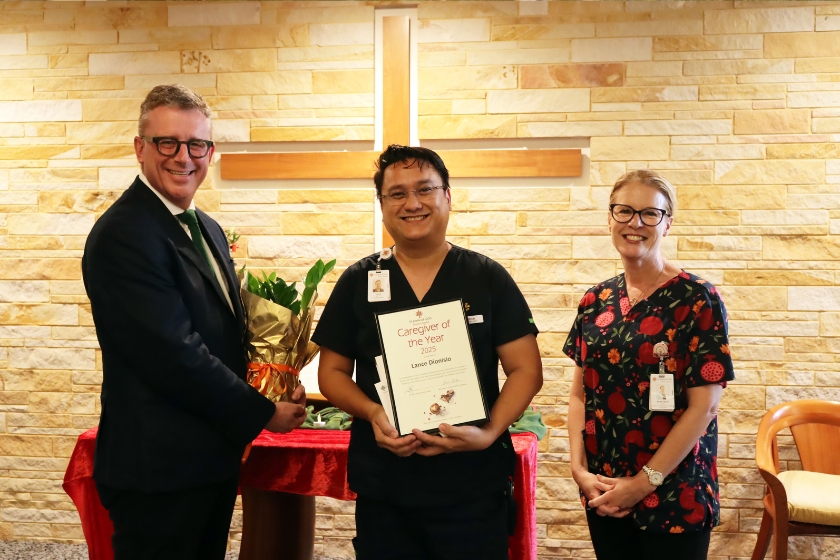 3 people are posing in front of the camera with a cross behind them, the middle person is holding a certificate