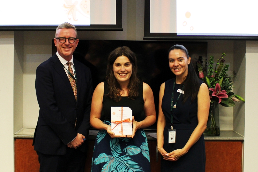 Three people at a formal event, with the person in the center holding a wrapped award while all smile for the photo.