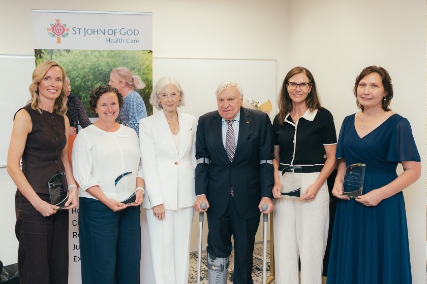 Six adults standing in a row at an awards presentation in front of a St John of God Health Care banner. Five women and one older man pose for a photo while holding glass award trophies.