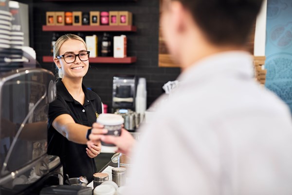 A female barrista with glasses hands a coffee to a male customer from behind the counter