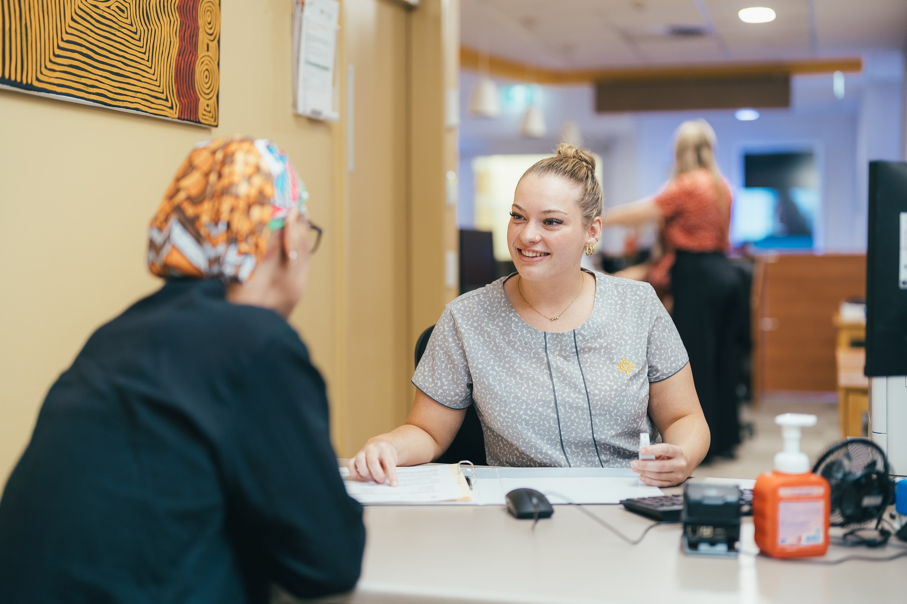 A female nurse in a grey scrub looks and smiles at a women sitting in front of her in a hospital