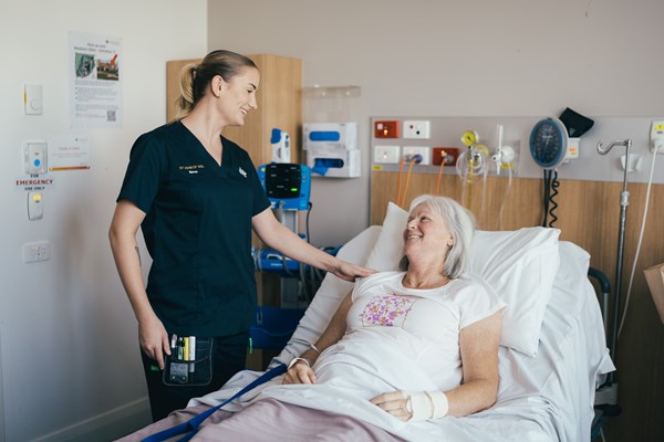 A nurse in a dark blue scrub caringly puts her hand on the shoulder of an elderly patient who is lying in a hospital bed, both looking at each other and smiling