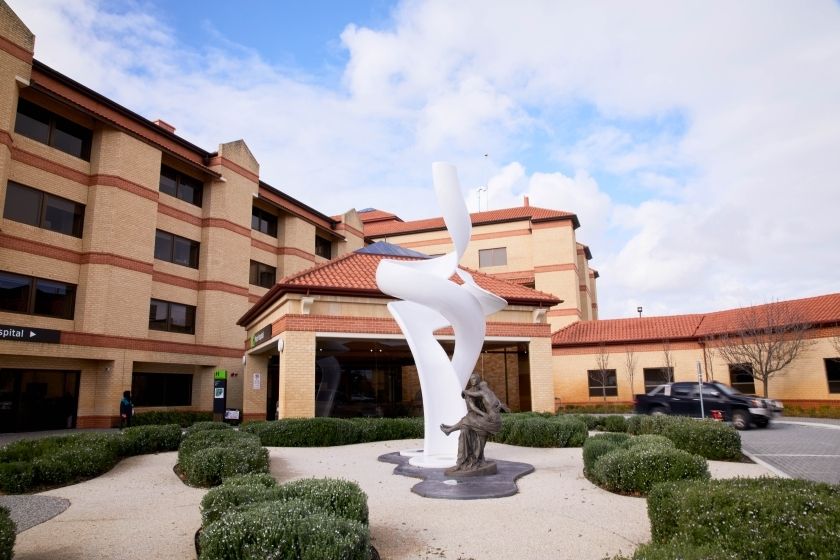 a picture of the entry to St John of God Murdoch, featuring a white abstract sculpture in front of the main entrance of a yellow and red building