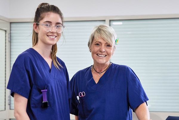 Two women in blue scrubs are smiling into the camera in front of a window with the blinds closed