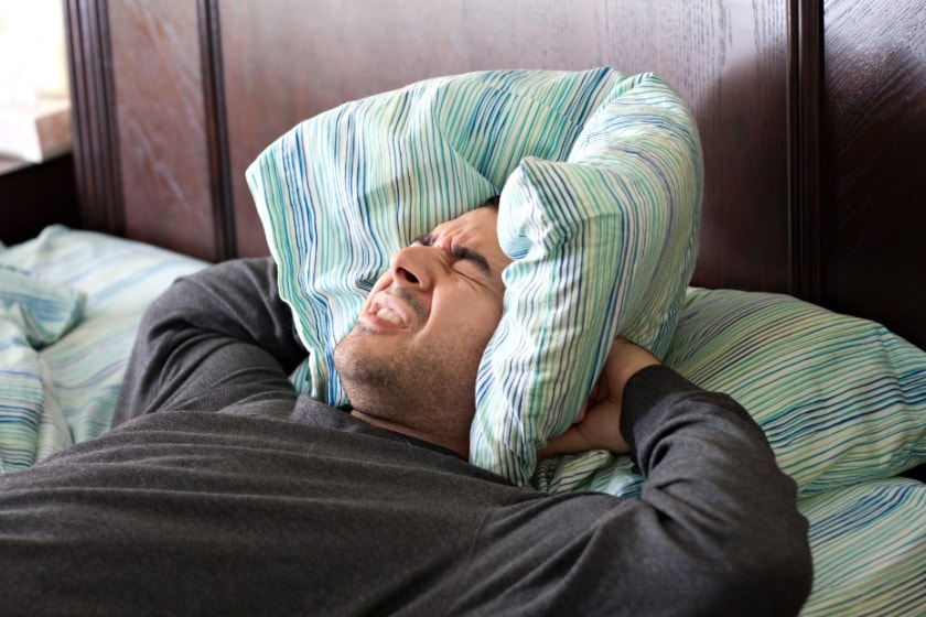 A man lying in bed pressing his cushion over his ears with an annoyed expression on his face