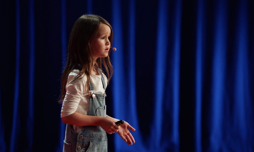 Seven year old Molly Wright on stage during her TED Talk with a blue curtain backdrop.