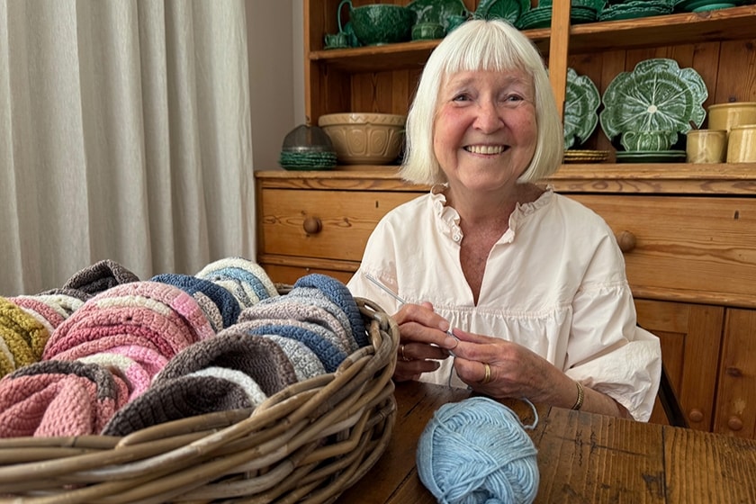 Joan Martin, an elderly lady sitting behind a table and smiling at the camera. She is busy crocheting with a blue yarn of wool, a basket of finished woolen hats is standing next to her.