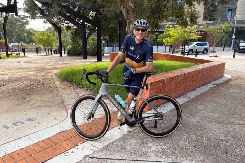 A man in a dark blye lycra cycling outfit, wearing a helmet and sunglasses stands behind his racebike with his elbow leaning on the saddle