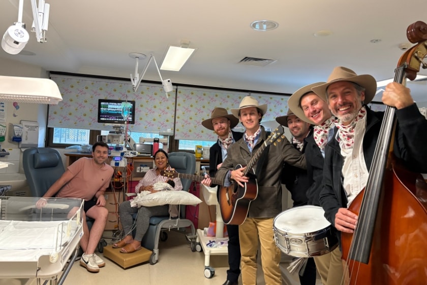 Five man with cowboyhats and instruments are standing in a hospital room with two parents and a baby, who is attached to a heart monitor, in the back. Everyone is smiling and the band is holding a drum, guitars and a cello.