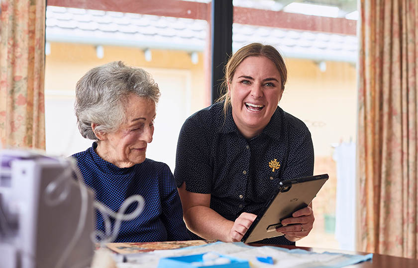 A caregiver holding an iPad smiling at the camera as they help a patient in their home.