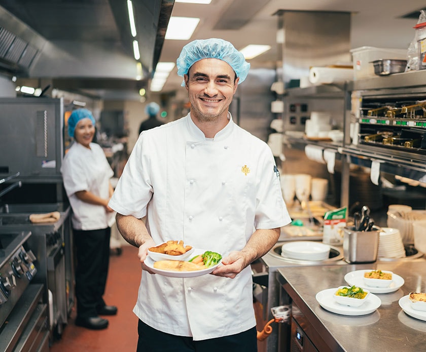 A catering caregiver in the kitchen smiles holding a freshly prepared meal