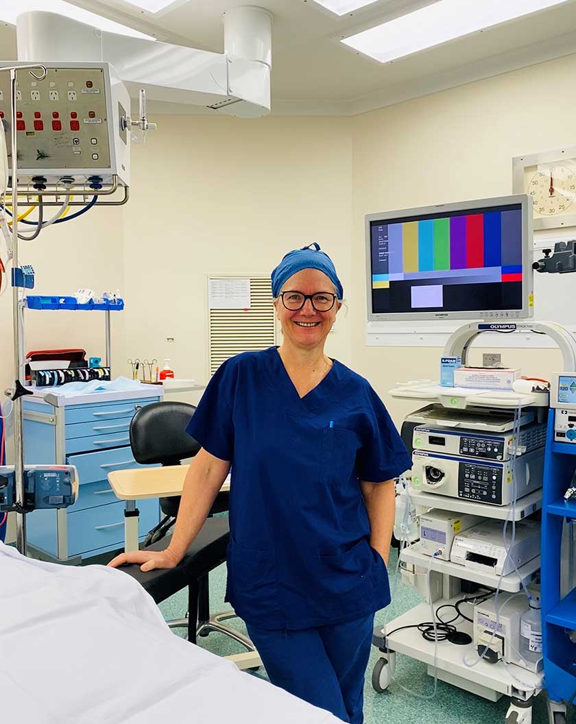 Dr Sue Chapman smiles for a photo in a theatre dressed in blue surgery scrubs.