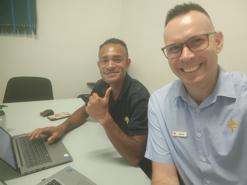 2 people sitting behing a desk working on their laptop, the one on the front taking a selfie and smiling at the camera with the person in the back raising his thumb
