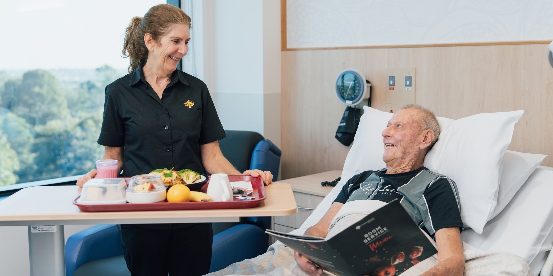 A caregiver carries a tray of food, smiling at a patient lying in bed, whilst the patient smiles back, holding a room service menu booklet.