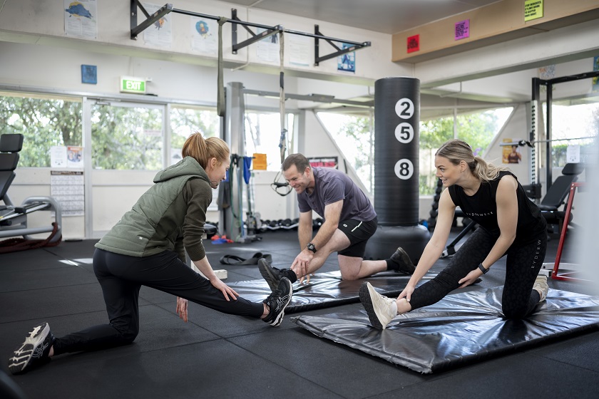 Trainer and patients stretching their legs on a mat in a small gymnasium