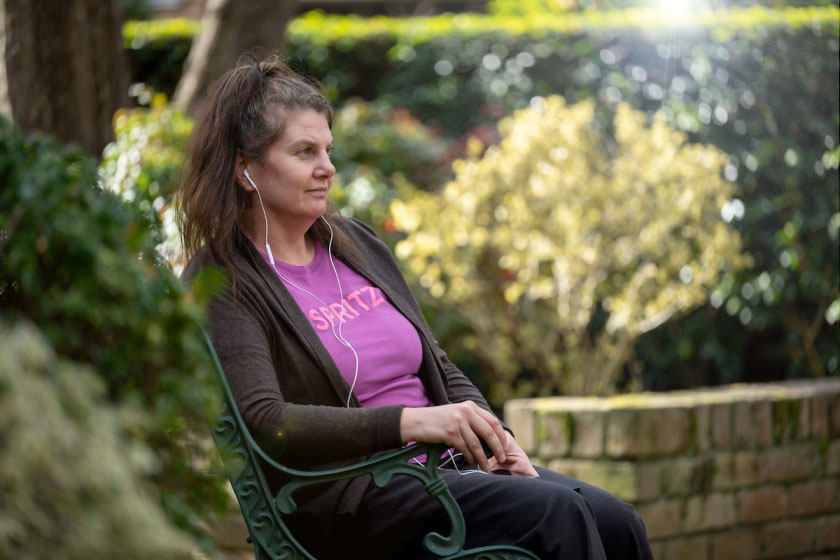 Person sitting on a park bench wearing wired earphones in an outdoor garden setting amongst trees and hedged gardens looking outwards.