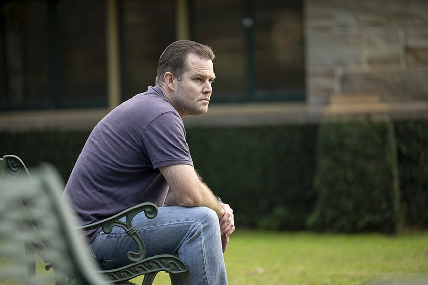 A person sits on a bench chair in an outdoor garden, looking off into the distance.