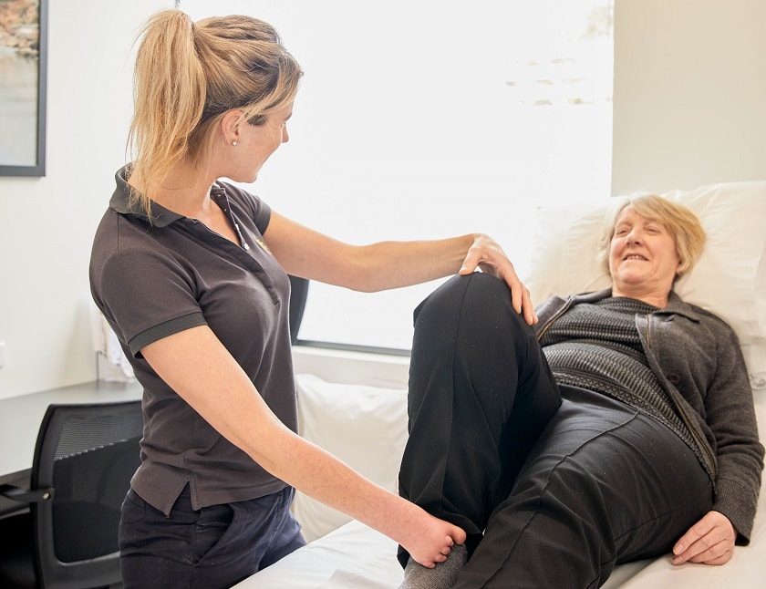 Allied health worker assists patient to move their knee while laying down on hospital bed