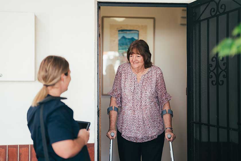 A lady in crutches stands talking with a caregiver at her front door